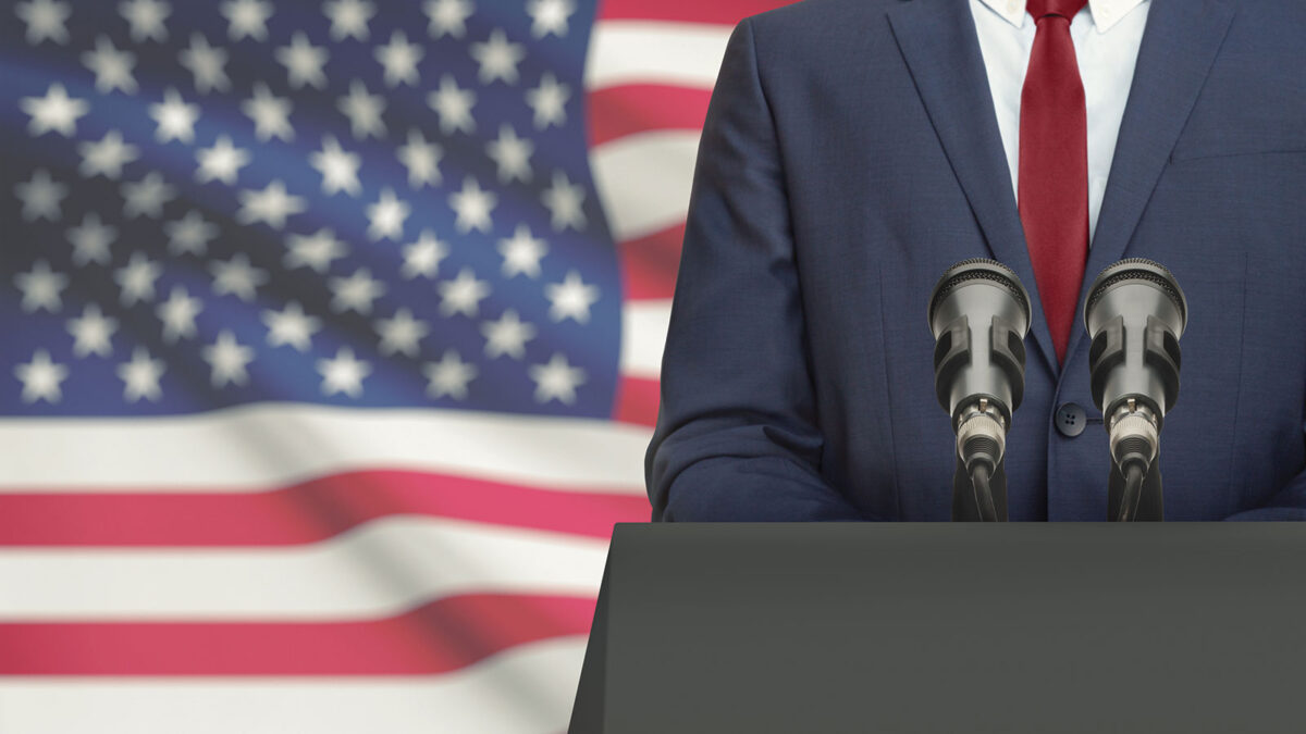 Man in suit at podium with American flag in background